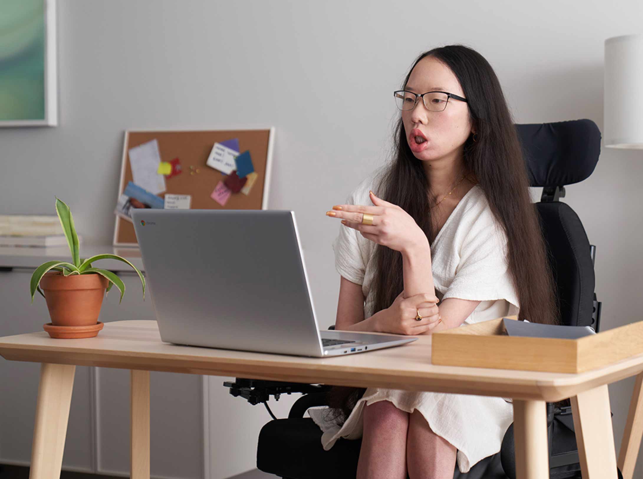 Jeune femme aux cheveux longs et portant des lunettes, assise à un bureau dans un fauteuil roulant électrique et regardant un Chromebook ouvert.